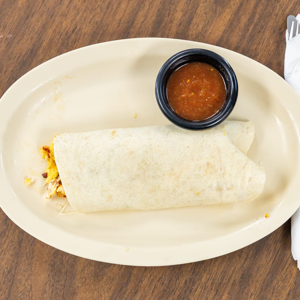 Breakfast burrito on a plate with scrambled eggs and fillings, served alongside a bowl of red salsa and wrapped utensils.