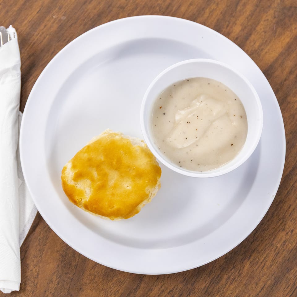 Golden biscuit with a bowl of peppered white gravy on a plate, accompanied by a napkin-wrapped fork on a wooden table.