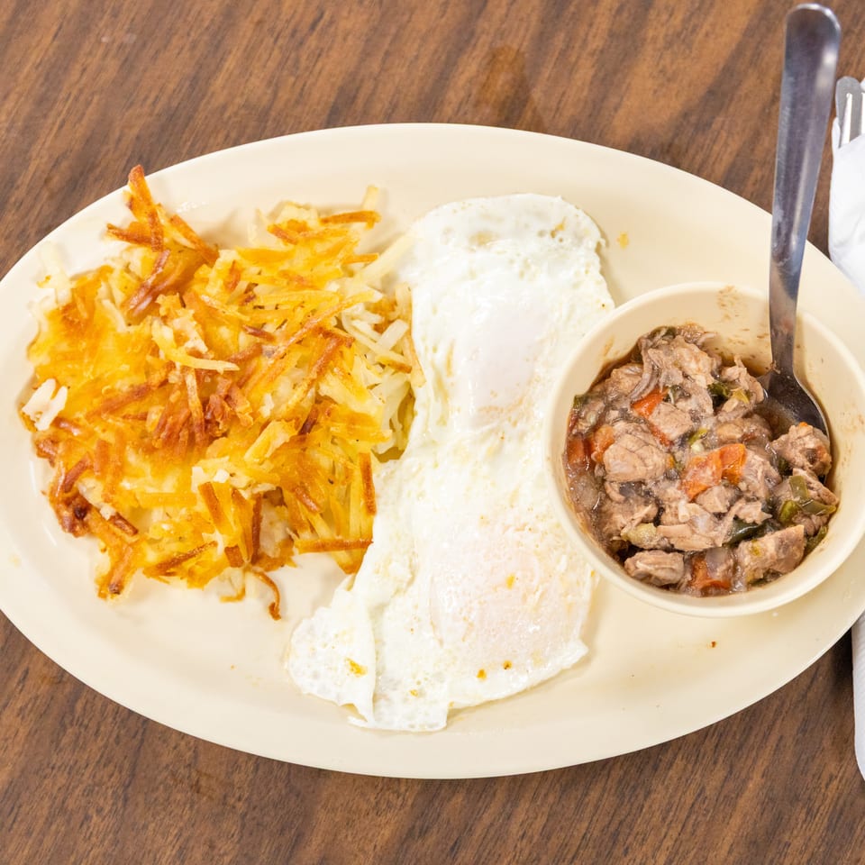 Plate of crispy hash browns, fried eggs, and a bowl of beef stew on a wooden restaurant table.