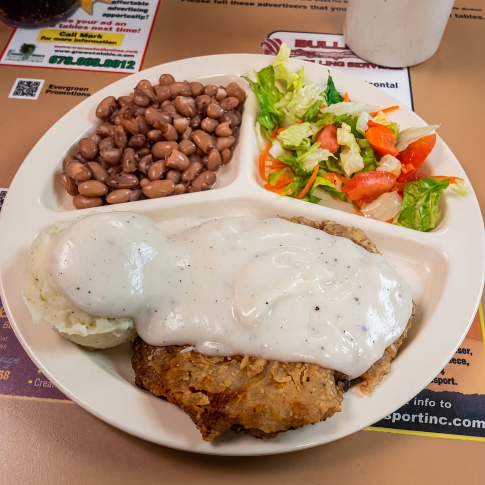 Plate of country-fried steak covered in white gravy, served with mashed potatoes, pinto beans, and a fresh salad.