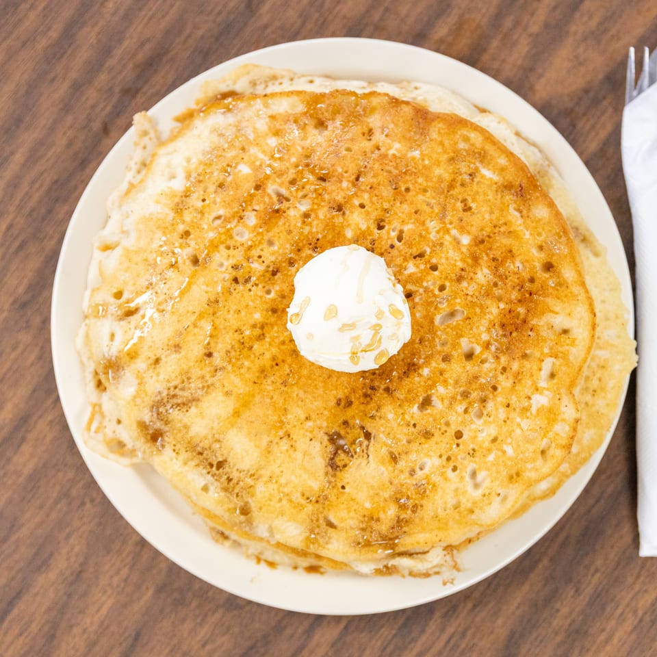 Stack of golden pancakes topped with butter and drizzled syrup on a white plate, with a napkin and utensils nearby.