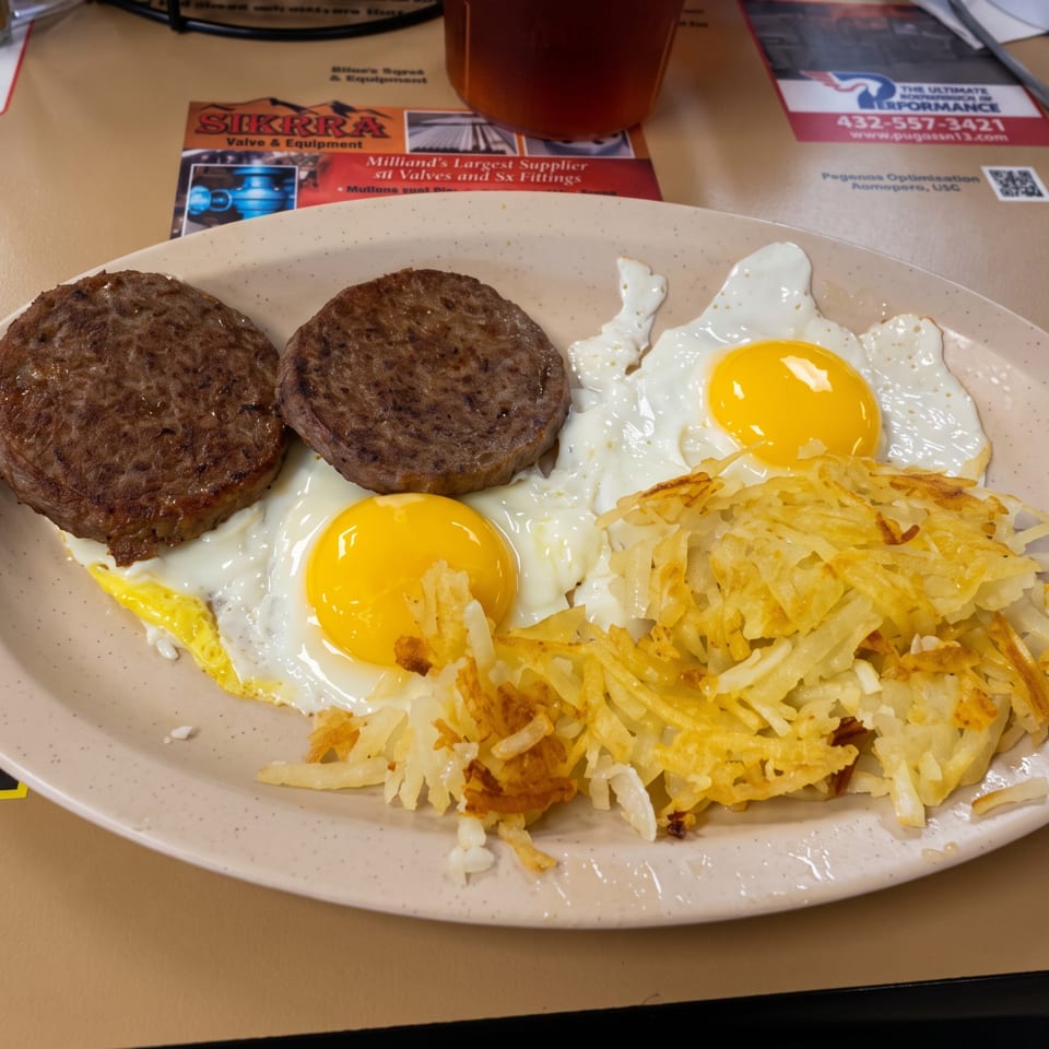 Breakfast plate with two sausage patties, two sunny-side-up eggs, and crispy shredded hash browns on a diner table.