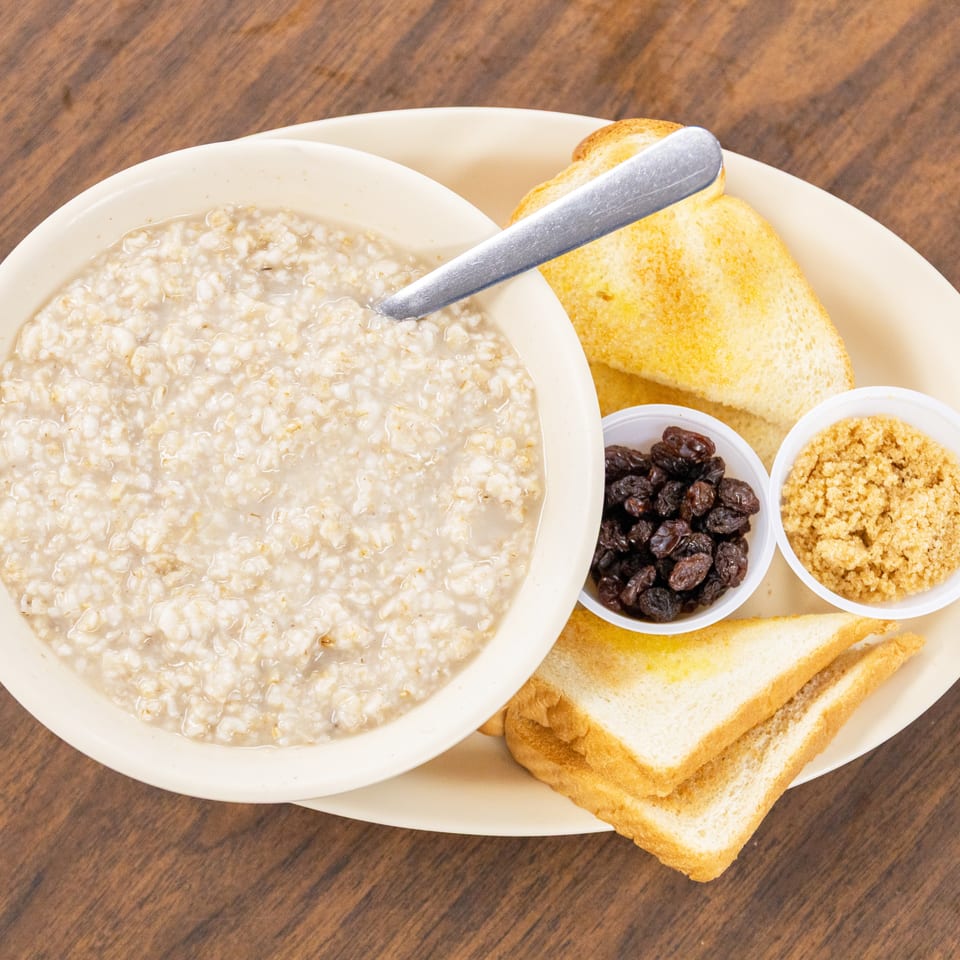 Bowl of oatmeal with toast, raisins, and brown sugar served on a plate over a wooden table.