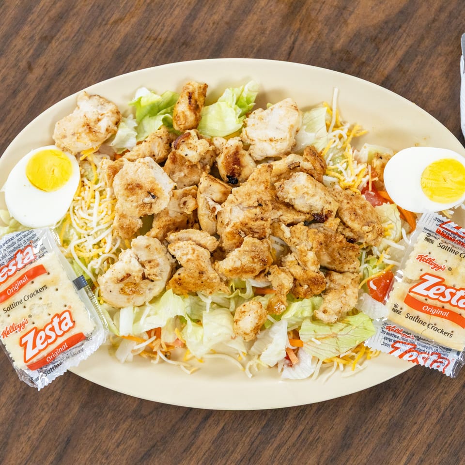 Large salad topped with grilled chicken, shredded cheese, vegetables, and halved boiled eggs, served with packets of Zesta crackers.